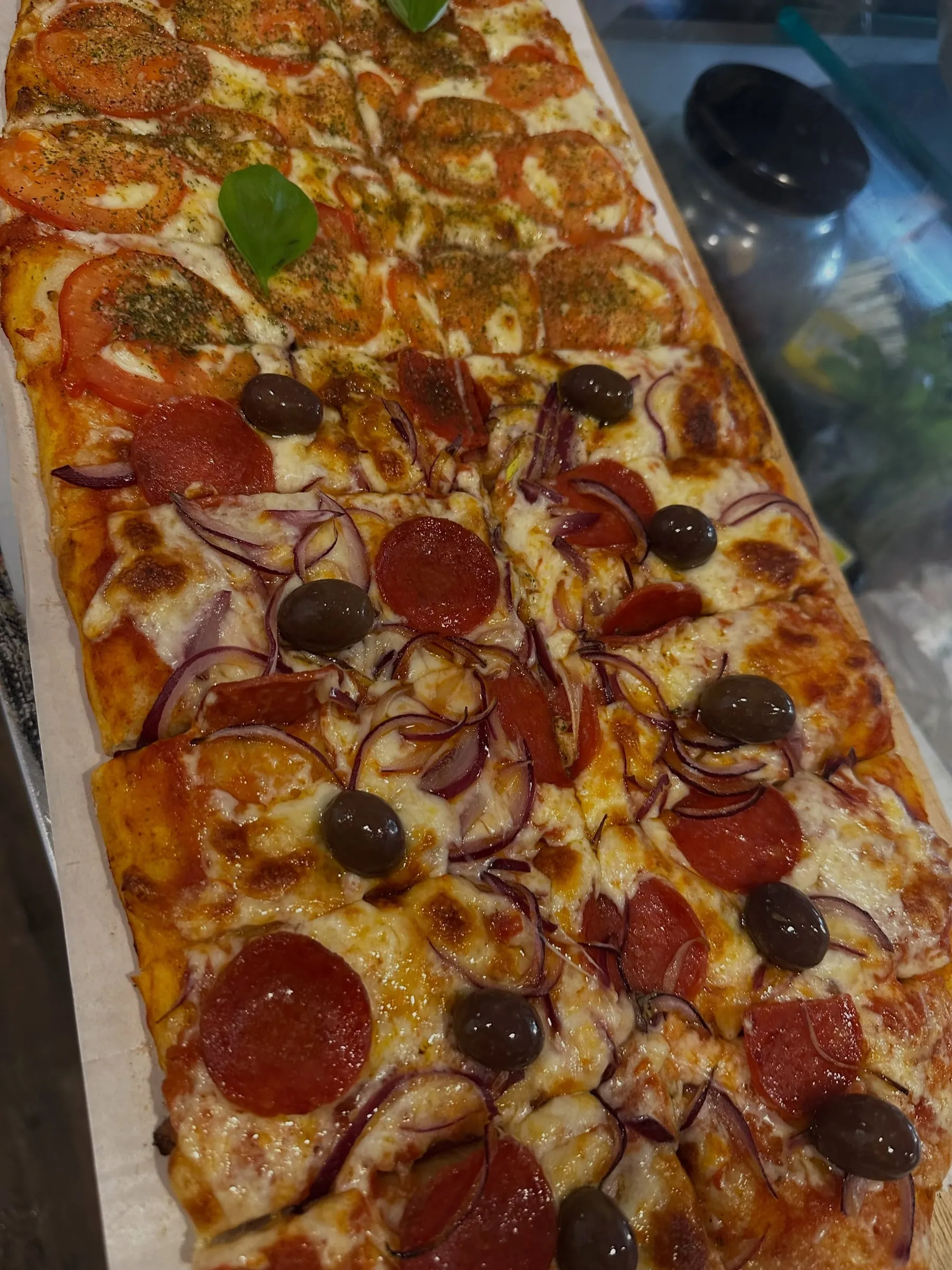 Uma pizza ao metro na vitrine, numa tábua comprida: tomate e manjericão numa ponta, pepperoni, cebola roxa e azeitonas na outra.
